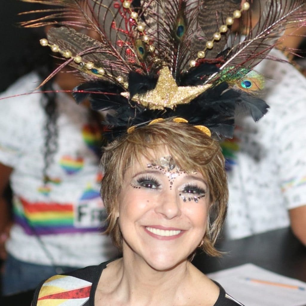 Uma pessoa sorridente usando maquiagem elaborada e um cocar de penas de pavão, detalhes dourados e um pássaro dourado está em frente a pessoas com camisetas com temas de arco-íris, capturando o espírito vibrante perto do Apartamento Lourdes bh.