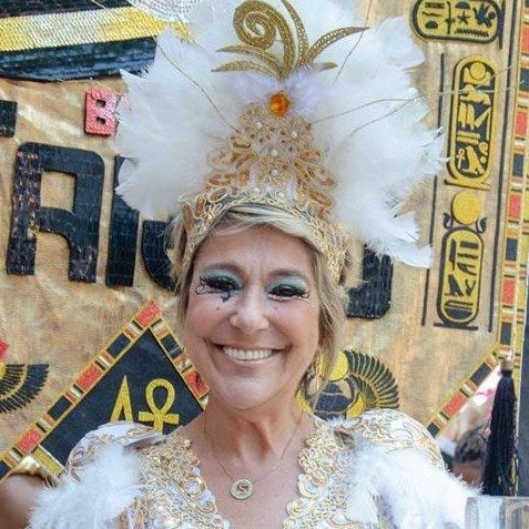 Uma mulher em um traje elaborado em dourado e branco, com um cocar de penas, sorri em frente a um cenário decorado com símbolos e hieróglifos de estilo egípcio no Apartamento Lourdes BH.