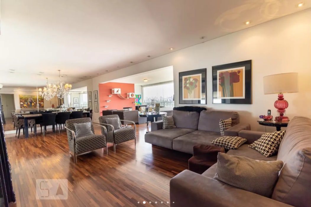 Ampla sala de estar e jantar com piso de madeira, sofá cinza moderno, poltronas estampadas, luminária vermelha, arte na parede, lustre e uma parede com detalhes em laranja ao fundo. Janelas amplas proporcionam luz natural.