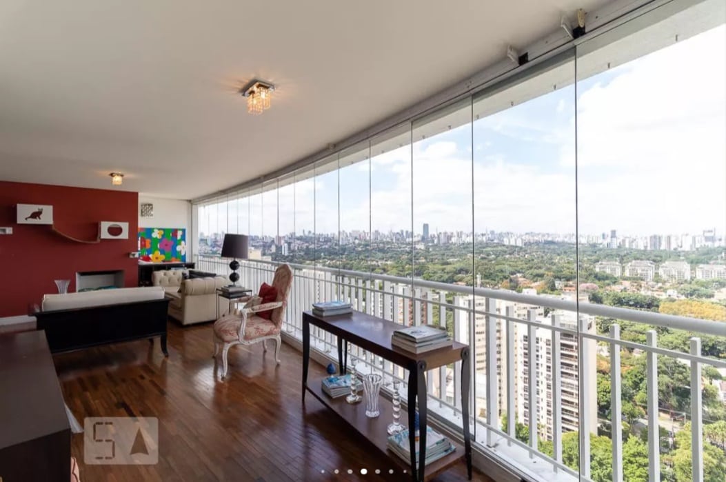 Sala de estar espaçosa e moderna com piso de madeira, parede com detalhes em vermelho e grandes janelas de vidro que oferecem vista panorâmica da cidade e do parque a partir de uma varanda no alto. Móveis e obras de arte contemporâneos estão expostos.