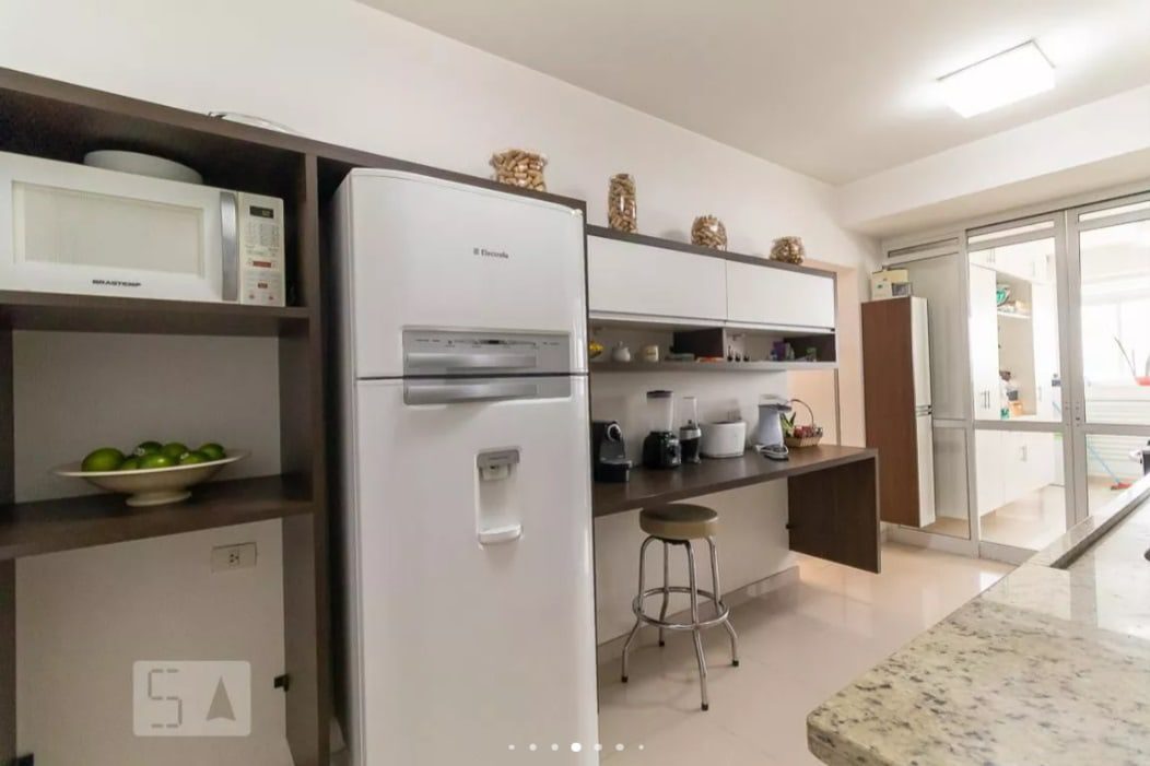 Cozinha moderna com geladeira branca, micro-ondas e prateleiras escuras. Bancada com pequenos eletrodomésticos e banquetas; portas de vidro ao fundo permitem a entrada de luz natural. Potes decorativos expostos nos armários superiores.