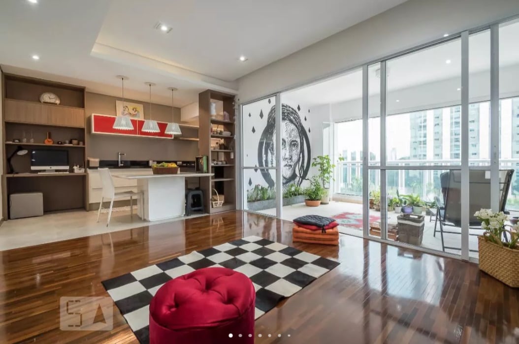 Sala de estar moderna em plano aberto com piso de madeira, tapete xadrez preto e branco, pufe vermelho, escrivaninha embutida, cozinha com luminárias pendentes, varanda com plantas e uma grande arte de parede com o rosto de uma mulher.