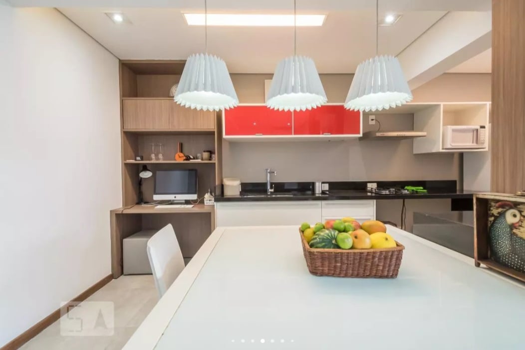 Uma cozinha e área de jantar modernas com uma mesa branca, uma cesta de frutas variadas, luminárias pendentes, armários vermelhos e de madeira, um micro-ondas e uma coruja decorativa em uma prateleira.