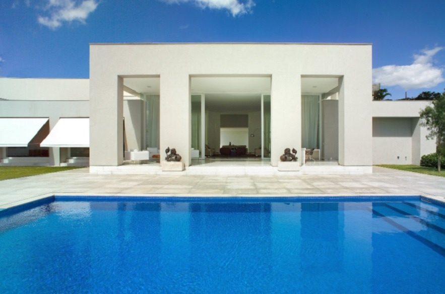 Casa branca moderna com grandes portas de vidro e arquitetura minimalista, vista através de uma piscina azul-clara; esculturas ficam ao lado da entrada — uma das elegantes Casas dos sonhos de BH sob um céu claro e brilhante.