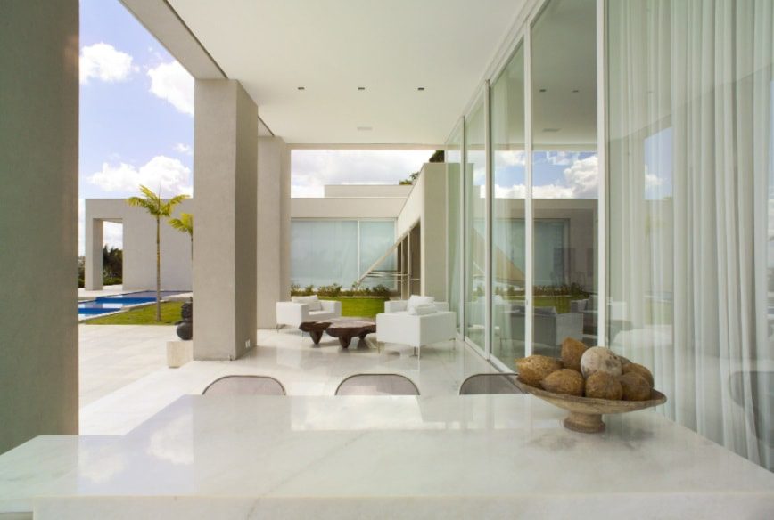 Área de pátio externa moderna com mesa de mármore branco, tigela decorativa, paredes de vidro e cadeiras brancas com vista para a piscina e o gramado sob um céu parcialmente nublado — um ambiente elegante e minimalista inspirado nas Casas dos sonhos de BH.