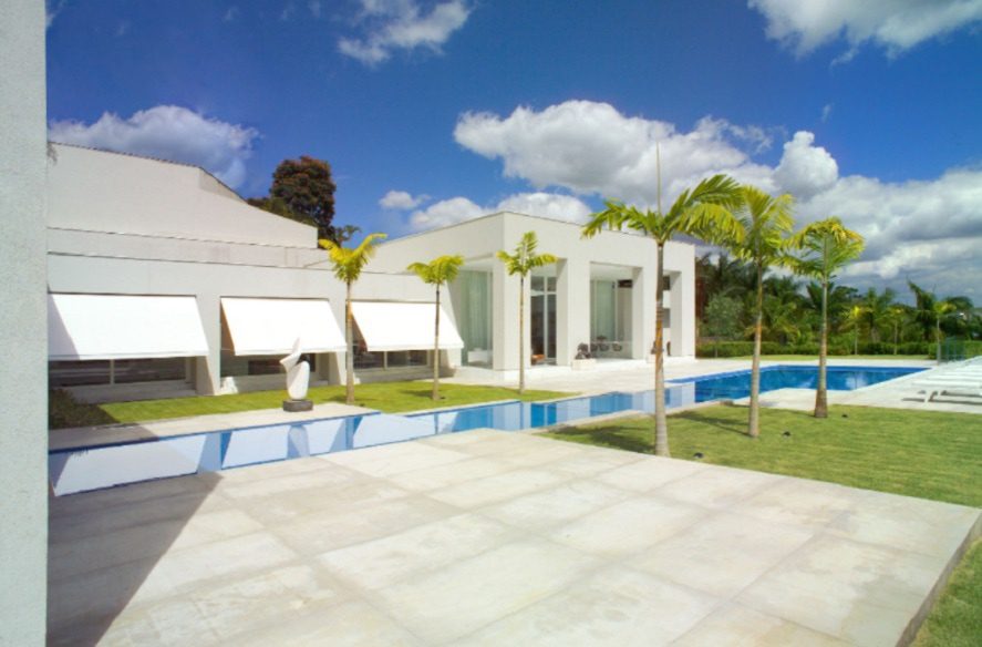 Uma moderna casa branca com grandes janelas e toldos dá para uma longa piscina, cercada por palmeiras e um gramado verde — um verdadeiro exemplo de Casas dos sonhos de BH — sob um céu azul brilhante com nuvens esparsas.
