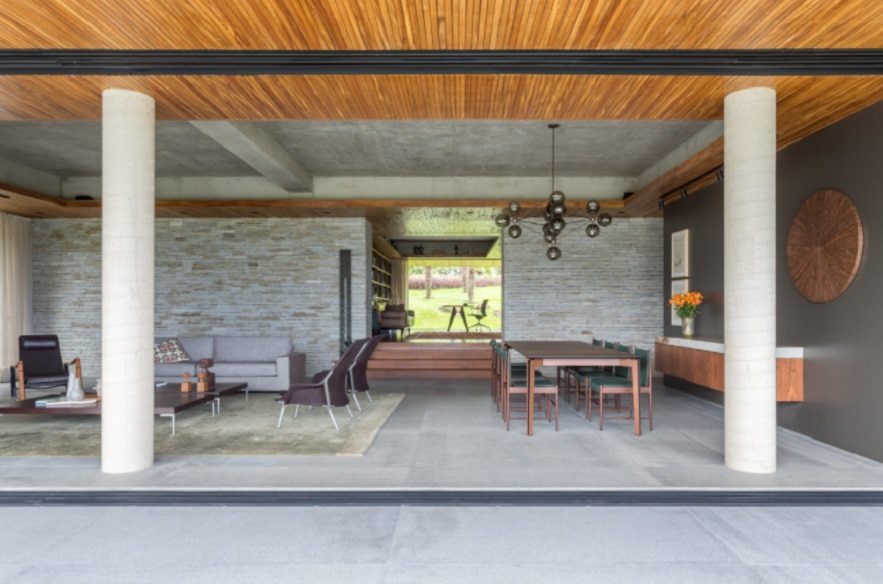 Uma moderna sala de estar e jantar em plano aberto com colunas de concreto, teto de madeira, móveis contemporâneos, uma mesa de jantar com cadeiras e uma grande janela com vista para o verde — perfeita para os admiradores das Casas dos sonhos de BH.
