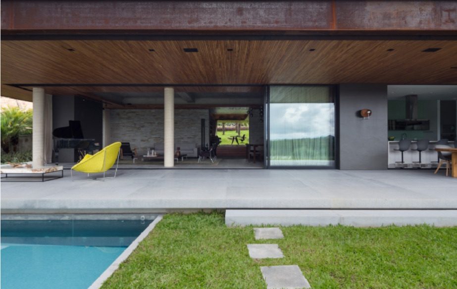 Casa moderna com sala de estar aberta, grandes portas de correr de vidro, cadeira amarela, teto de madeira e cozinha à vista. Uma piscina e um caminho de pedra levam ao quintal gramado — uma das Casas dos Sonhos de BH.