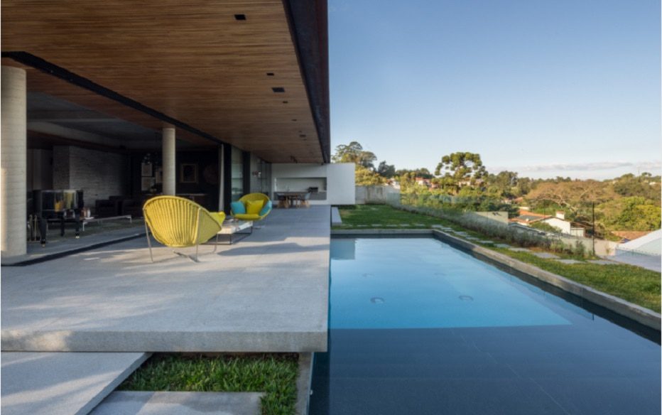Uma casa moderna com uma área de estar aberta, cadeiras amarelas ao ar livre e uma elegante piscina retangular com vista para uma paisagem verde — uma das verdadeiras Casas dos sonhos de BH sob um céu limpo.