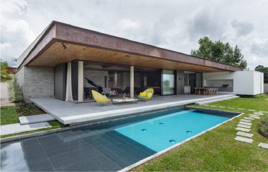 Casa térrea moderna com telhado plano, pátio aberto e paredes de vidro do chão ao teto — uma das Casas dos Sonhos de BH. Uma piscina retangular fica na frente, com mesas ao ar livre e cadeiras amarelas no terraço coberto.