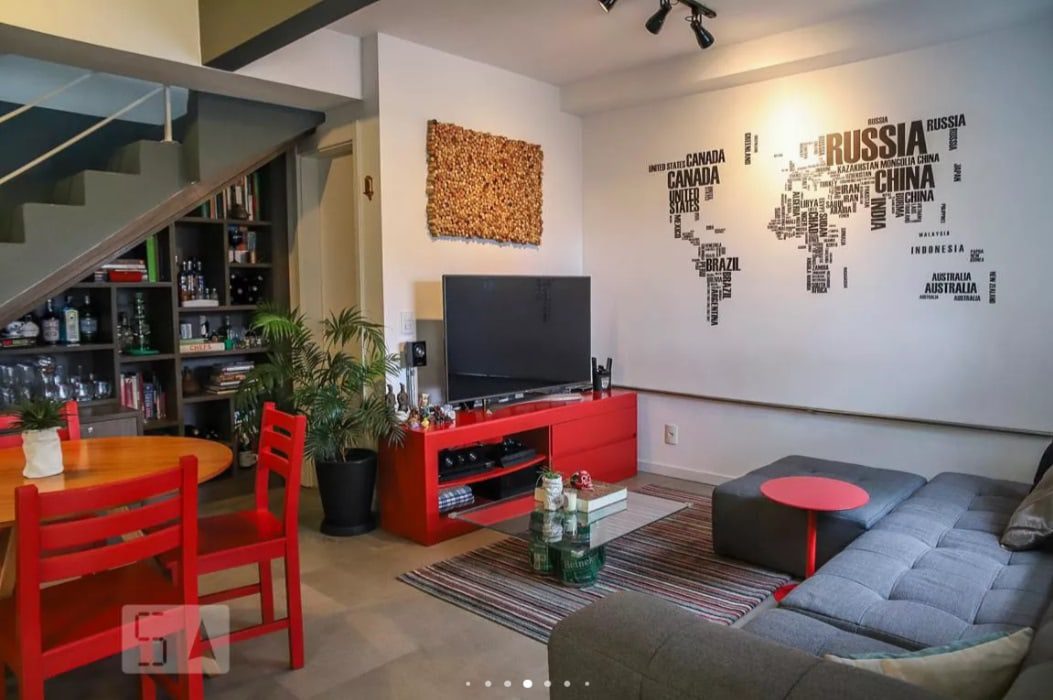 Sala de estar moderna com sofá cinza, suporte de TV e mesa vermelhos, TV na parede, arte com mapa-múndi, estante embaixo da escada, plantas de interior e um tapete listrado colorido. As escadas levam para cima pelo lado esquerdo.
