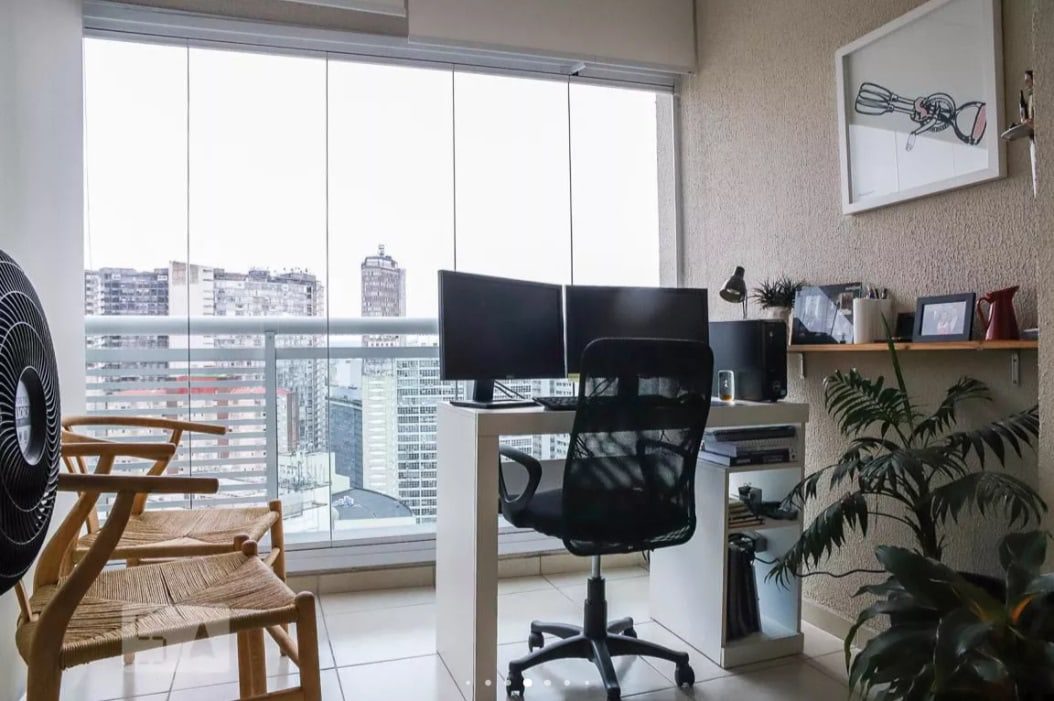 Um home office moderno com mesa, dois monitores, cadeira de escritório e prateleiras, situado ao lado de uma grande janela com vista para a paisagem urbana. Há uma planta, uma obra de arte na parede e cadeiras adicionais no ambiente iluminado e organizado.