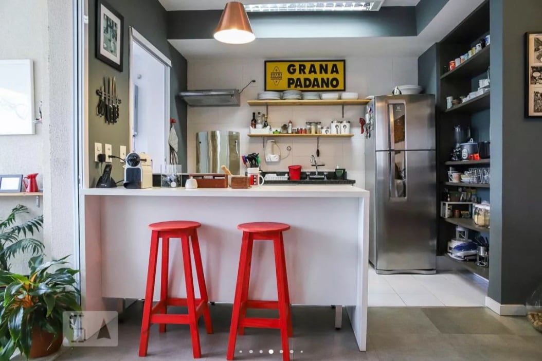 Cozinha moderna com ilha branca, duas banquetas vermelhas, geladeira de aço inoxidável, prateleiras abertas e uma placa amarela da Grana Padano na parede. Luminária pendente pendurada acima da ilha; plantas e utensílios de cozinha visíveis.