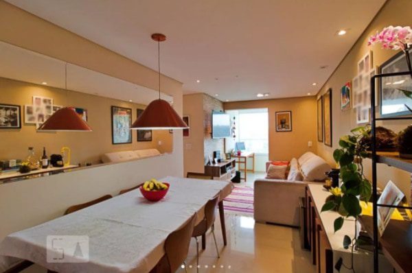 Uma sala de jantar e estar bem iluminada com mesa e cadeiras, luminárias pendentes vermelhas, um sofá, quadros, plantas, prateleiras e uma TV. Uma grande janela na extremidade permite a entrada de luz natural.