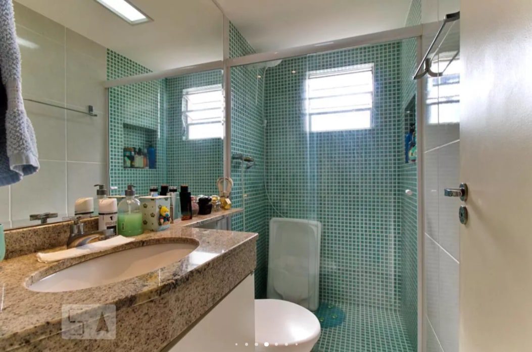 Banheiro moderno com paredes de mosaico verde no chuveiro, bancada de granito com vários produtos de higiene pessoal, uma toalha pendurada no lado esquerdo, um vaso sanitário e duas pequenas janelas que deixam entrar luz natural.