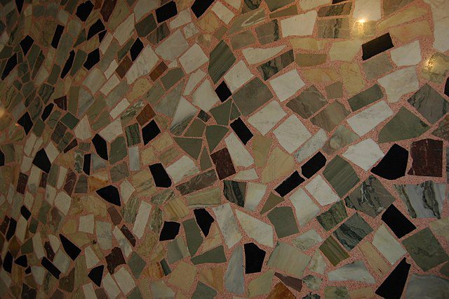 Uma parede de mosaico feita de azulejos de formas irregulares em verde, branco, marrom e preto com rejunte rosa claro evoca o charme aconchegante de uma Casa de vó, preenchendo o espaço com aconchego e um caráter nostálgico.