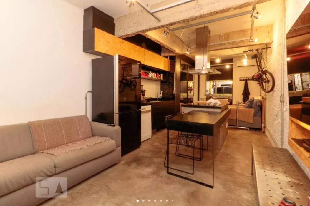 Apartamento moderno e aberto com piso de concreto, vigas aparentes, um sofá cinza, armários de cozinha pretos, uma ilha central com duas cadeiras e uma bicicleta pendurada na parede perto da entrada. Iluminação aconchegante em todo o ambiente.