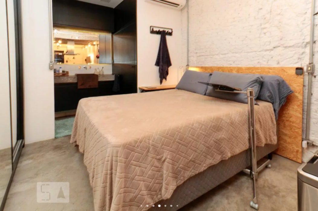 Um quarto moderno com uma cama de casal coberta com uma colcha bege, almofadas azuis e uma cabeceira de madeira. O quarto tem piso de concreto, uma parede de tijolos brancos, uma escrivaninha na parede e vista para um banheiro.