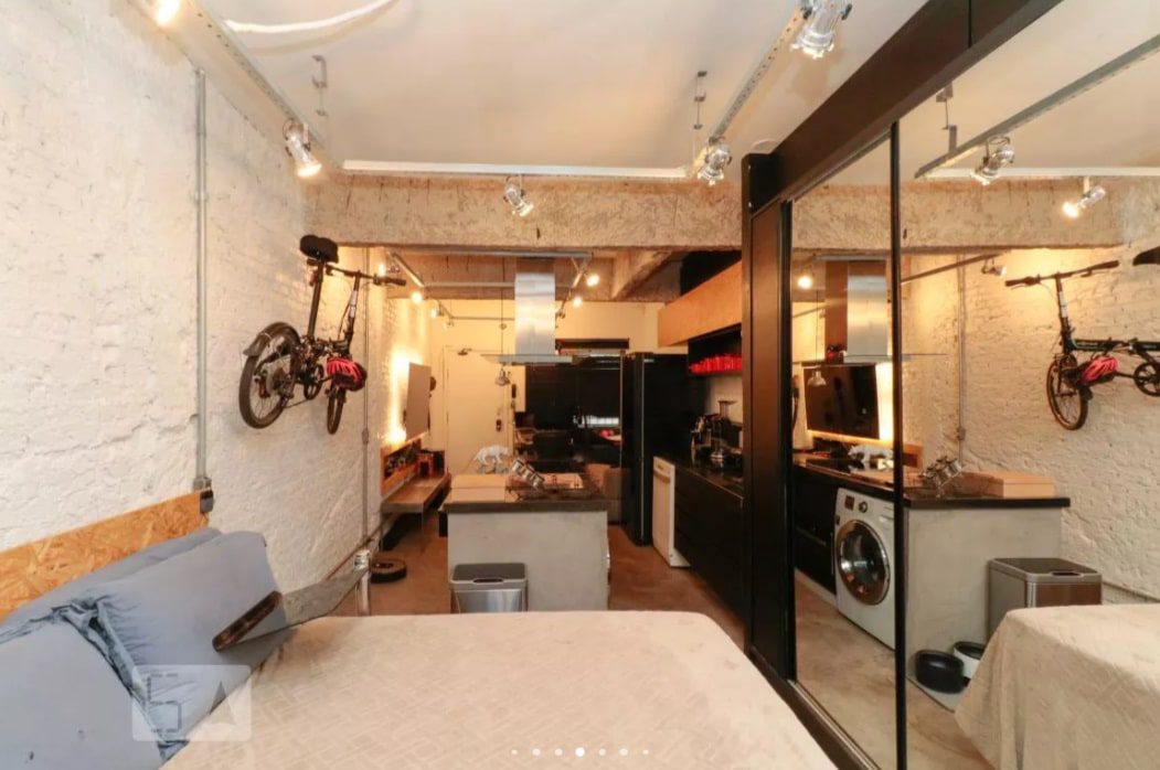Um apartamento estúdio moderno com paredes de tijolos brancos, uma cama no primeiro plano, uma pequena cozinha no fundo, uma bicicleta montada na parede, uma máquina de lavar e armários espelhados refletindo o espaço.