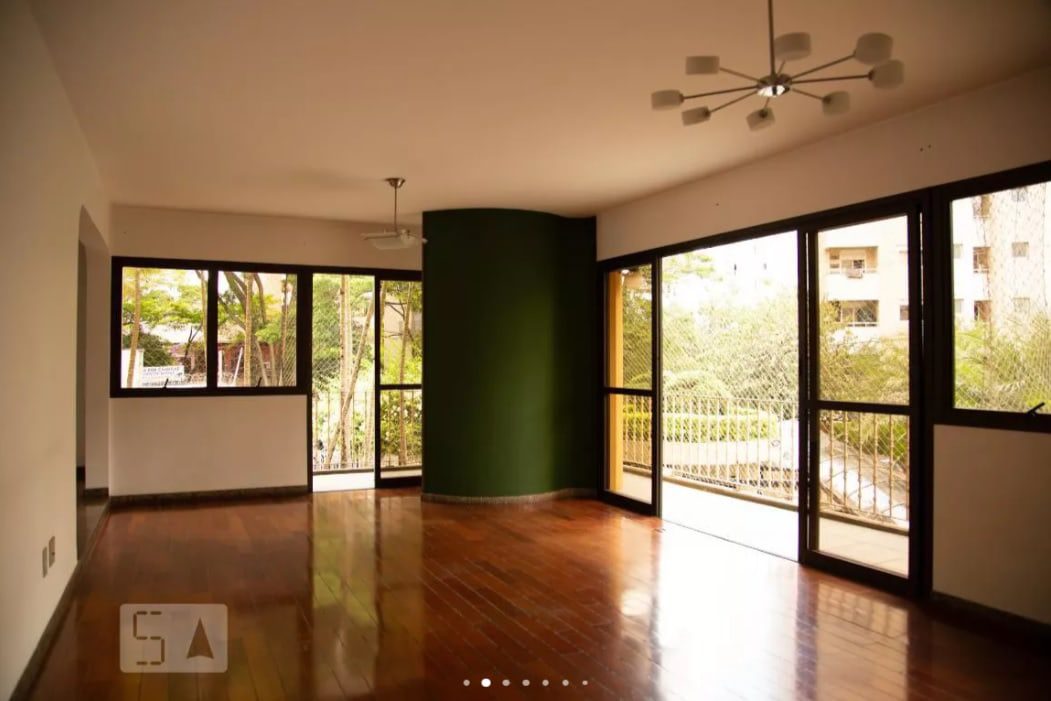 Sala de estar espaçosa com piso de madeira polida, janelas amplas e uma porta de correr de vidro que dá acesso a uma varanda. As paredes são predominantemente brancas, com uma parede verde-escura destacada, e duas luminárias modernas no teto são visíveis.