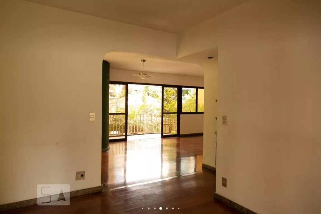 Uma sala iluminada e vazia, com piso de madeira polida, paredes brancas, janelas grandes e uma porta de vidro que dava para uma varanda com árvores verdes visíveis do lado de fora. Um ventilador de teto está pendurado na sala ao lado.