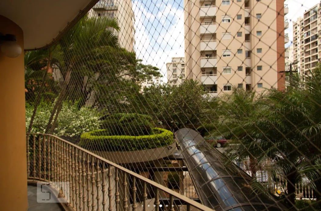 Vista de uma varanda com rede de segurança, com vista para um pátio paisagístico com arbustos, palmeiras e uma passarela coberta, cercado por altos prédios de apartamentos.