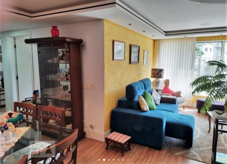 Uma sala de estar aconchegante com um sofá secional azul, almofadas coloridas, um abajur, quadros emoldurados em uma parede com detalhes em amarelo e um armário de vidro. Grandes janelas com persianas verticais permitem a entrada de luz natural; plantas e a mesa de jantar também estão visíveis.