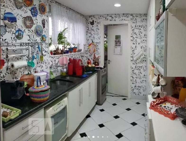 Uma cozinha aconchegante com armários brancos, bancadas pretas, piso de ladrilhos estampados em preto e branco e papel de parede floral. Pratos e panelas coloridas decoram as paredes e as bancadas. Uma janela com cortinas brancas permite a entrada de luz natural.