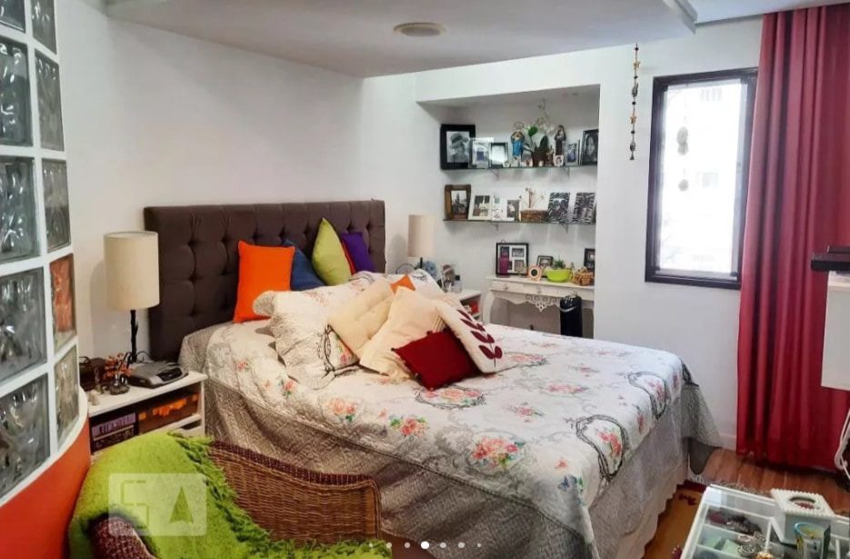 Um quarto aconchegante com uma cama de casal coberta por um edredom com estampa floral e almofadas decorativas. Há prateleiras com fotos e decoração, uma janela com cortinas vermelhas e vários itens pessoais sobre uma escrivaninha e uma mesa.