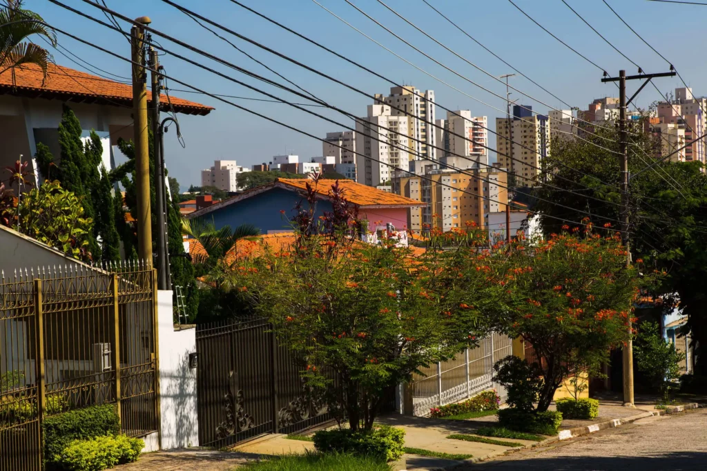 Imagem de rua residencial com casas fechadas, árvores verdes exuberantes e linhas de energia aéreas em primeiro plano no Alto da Lapa, em São Paulo. Prédios altos de apartamentos se erguem no fundo distante sob um céu azul claro.