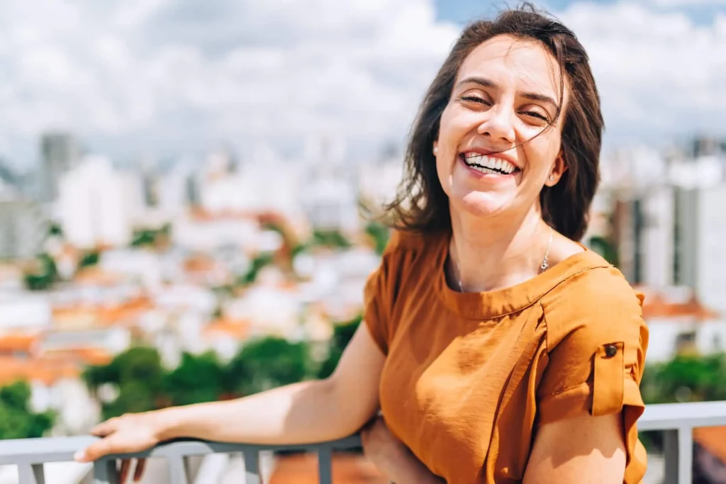 Imagem de uma mulher sorrindo em primeiro plano enquanto prédios estão ao fundo em segundo plano