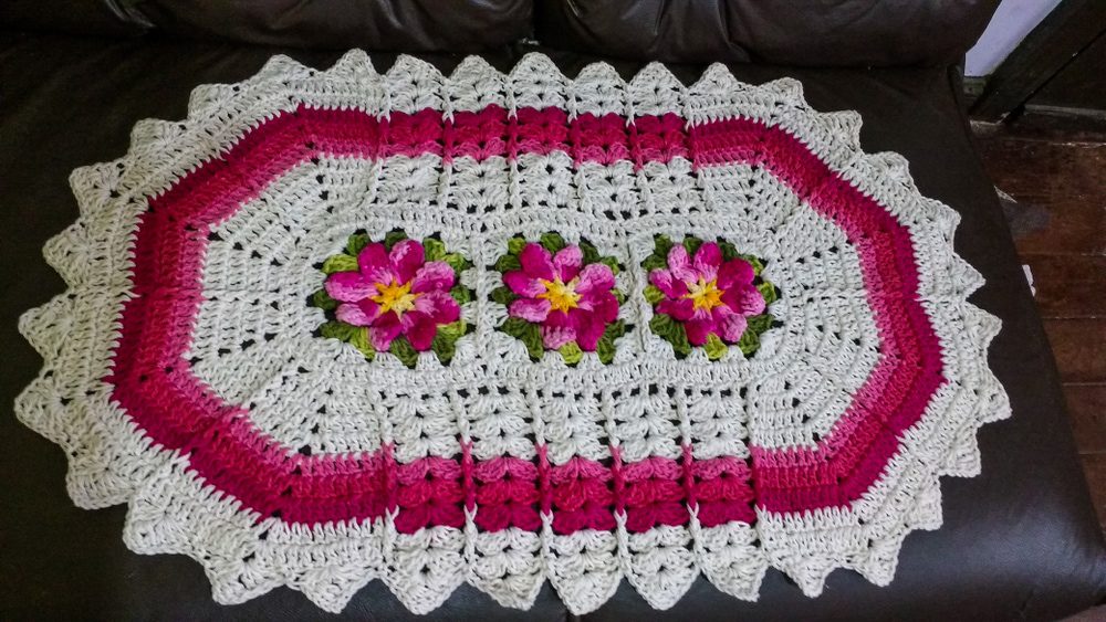 Um tapete oval de crochê com bordas recortadas, com base creme, bordas rosa escuro e três grandes motivos florais rosas e amarelos com folhas verdes no centro, evoca um clima aconchegante de Casa de vó em uma superfície escura.