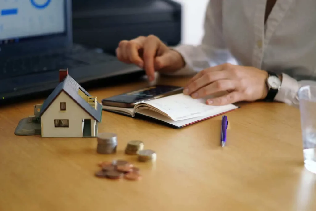 Imagem de um homem fazendo contas na calculadora de um celular próximo a um modelo de casa que está sobre a mesa para ilustrar matéria sobre como funciona a portabilidade de financiamento imobiliário