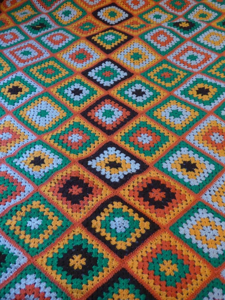 Uma manta de crochê colorida com um padrão repetido de quadrados da vovó em formato de diamante nas cores laranja, amarelo, verde, preto e branco. Lembrando uma Casa de Vó, os quadrados unidos criam um desenho geométrico vibrante.