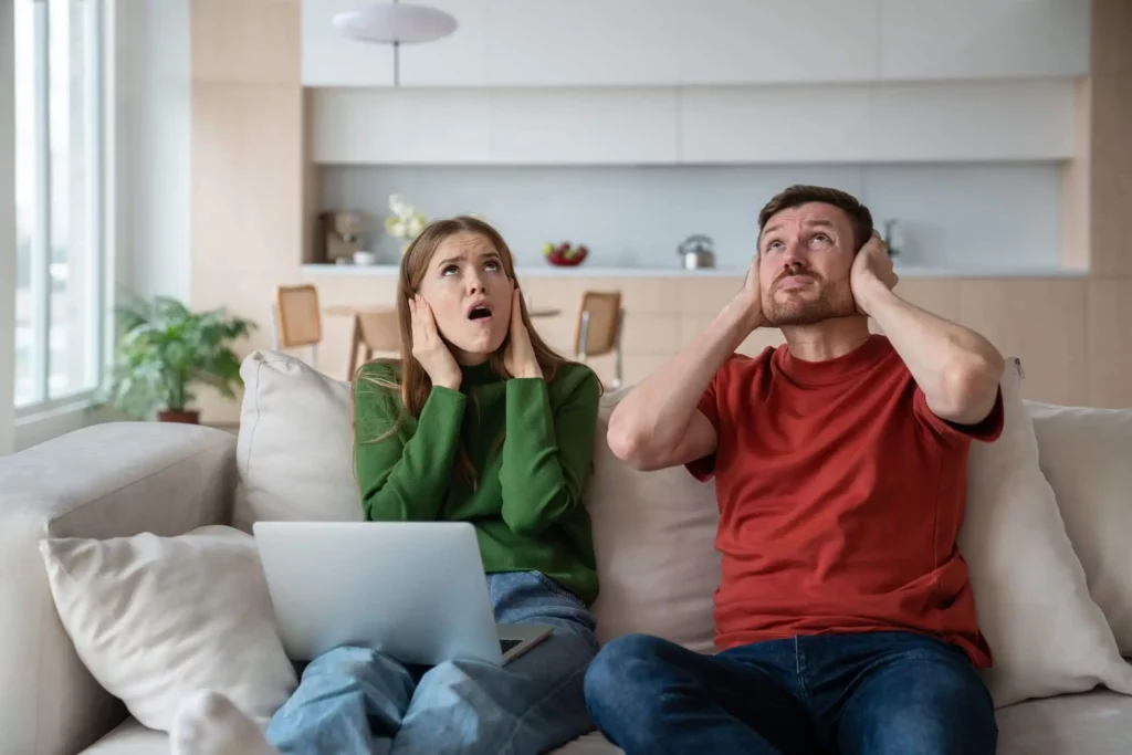 Imagem de um casal formado por um homem e uma mulher sentados no sofá de casa com as mãos nas orelhas, olhando para cima e com expressão de raiva para ilustrar matéria sobre a Lei do Silêncio