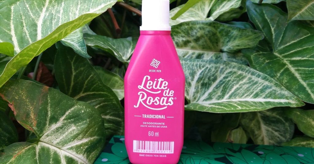 Um frasco rosa de 60 ml do desodorante Leite de Rosas se destaca entre grandes folhas verdes, evocando memórias da Casa de Vó. O rótulo branco exibe o nome da marca e as informações do produto em português.