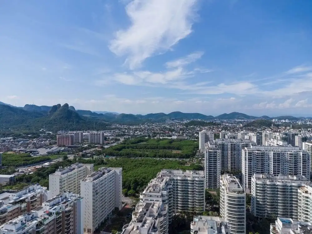 Foto que ilustra matéria sobre os melhores bairros do RJ para morar mostra uma vista aérea de Jacarepaguá, com grandes condomínios de prédios residenciais em primeiro plano e montanhas ao fundo em um dia de céu azul