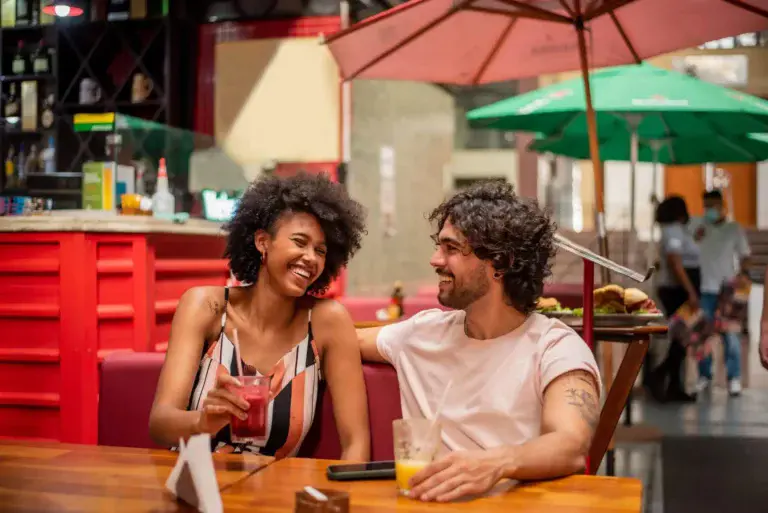 Imagem de um casal formado por um homem e uma mulher sentados na mesa de um restaurante enquanto conversam e sorriem para ilustrar matéria sobre os melhores bairros em São Paulo para morar