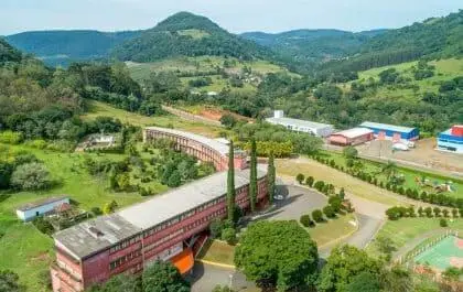 Imagem da vista aérea de um grande edifício de tijolos vermelhos cercado por árvores e vegetação, com montanhas e mais edifícios ao fundo, na cidade de Luzerna, em Santa Catarina