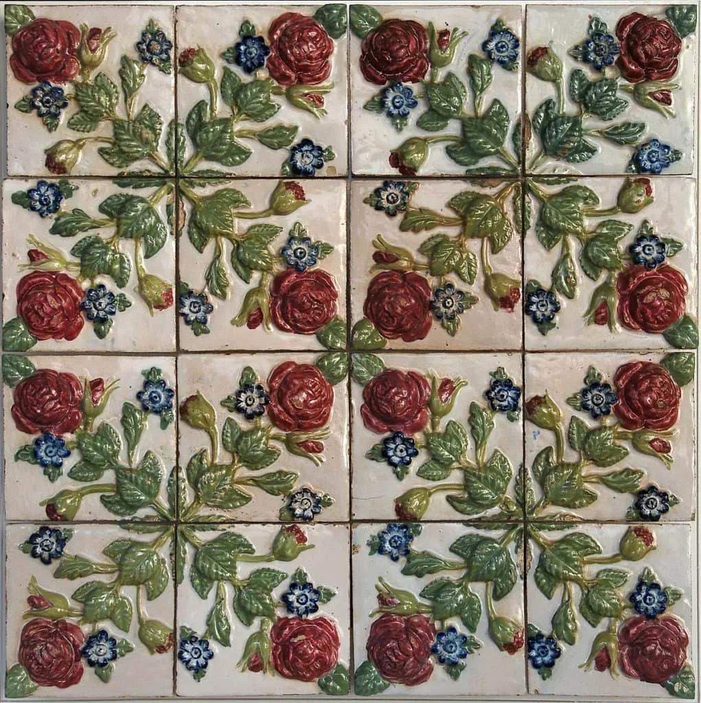 Azulejos cerâmicos decorativos dispostos em uma grade de 4x4, apresentando padrões florais em relevo com rosas vermelhas, folhas verdes e pequenas flores azuis sobre um fundo creme. O design simétrico evoca o charme aconchegante de uma Casa de Vó.