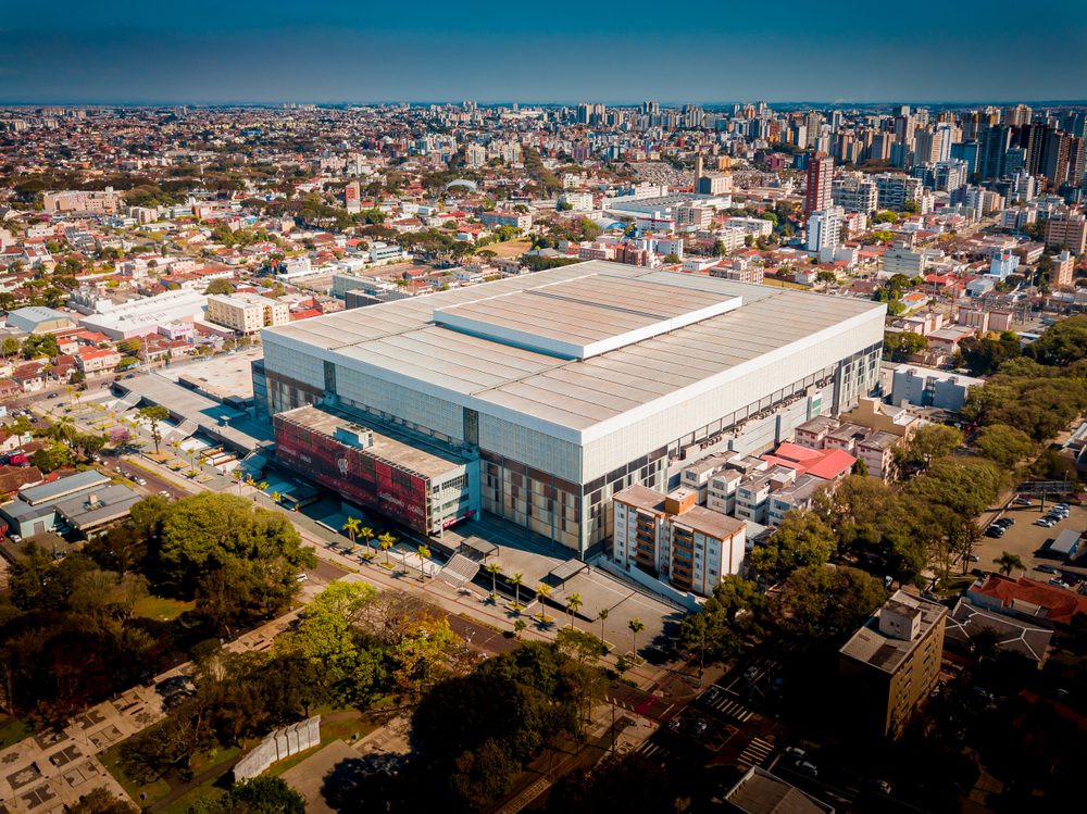 Vista aérea de uma grande e moderna arena cercada por árvores e prédios urbanos em um dos bairros nobres de Curitiba em um dia claro.