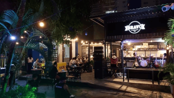 Vista externa de um animado restaurante-bar à noite em um dos melhores bairros de Salvador, com pessoas sentadas em mesas sob luzes de corda. Um pequeno palco com instrumentos musicais está montado perto da entrada, cercado por plantas tropicais.