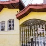 Uma pequena casa amarela com telhado de telhas vermelhas, janelas e portas arqueadas protegidas por barras de ferro brancas decorativas, e um número de casa 356 parcialmente visível. As paredes externas têm um pouco de tinta cinza escura perto do fundo.