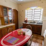 Uma cozinha com armários de madeira, uma mesa oval vermelha para quatro pessoas, cadeiras de metal, uma geladeira branca, fogão, filtro de água e uma janela com detalhes em vitral acima da pia.