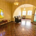 Uma sala de estar espaçosa com piso de madeira estampado, porta em arco, lustres vintage, vitrais, um sofá, uma pequena mesa de madeira e uma porta de entrada com vitrais coloridos.