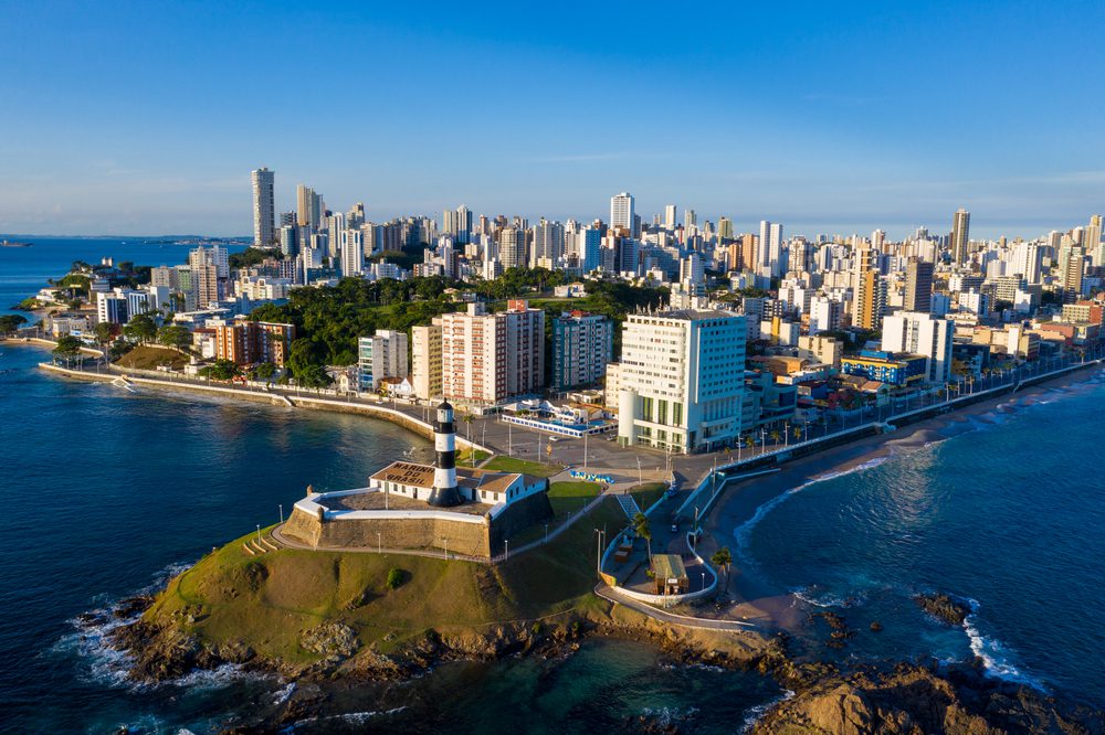 Vista aérea de uma cidade costeira com um farol proeminente em um ponto gramado, oceano azul, arranha-céus modernos e um céu claro, mostrando um dos melhores bairros de Salvador.