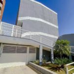 Edifício cinza moderno com linhas geométricas, grades brancas, porta de garagem fechada e pequenas palmeiras perto da entrada, sob um céu azul claro.