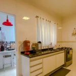 Uma cozinha iluminada com paredes brancas, bancadas pretas e um fogão. Uma janela com cortinas brancas permite a entrada de luz natural. O cômodo ao lado tem um pendente vermelho e uma pequena mesa com banquetas.
