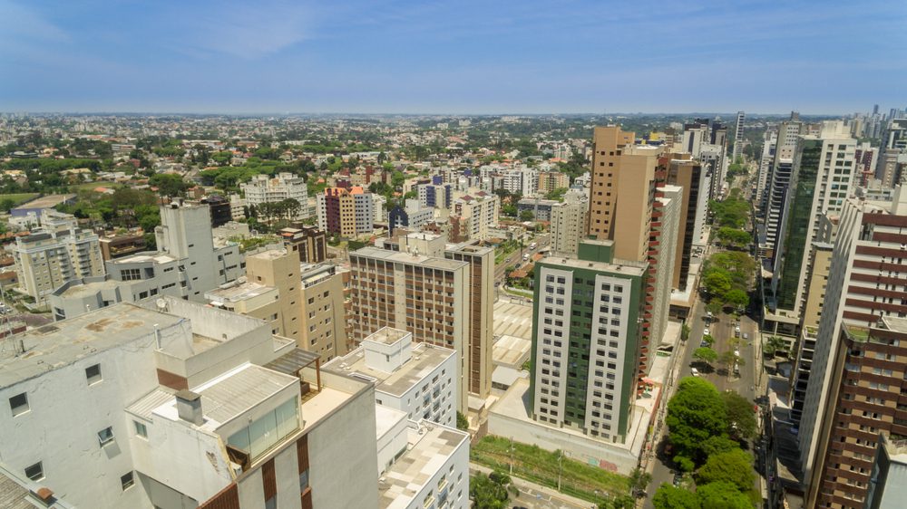 Vista aérea dos bairros nobres de Curitiba, com prédios altos e modernos, complexos de apartamentos e ruas arborizadas sob um céu azul que se estende até a distância.