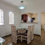 Uma sala de jantar com uma mesa de vidro e seis cadeiras de madeira fica ao lado da cozinha. O cômodo tem janelas em arco, piso de cerâmica e uma luminária pendente acima da mesa. A cozinha é visível através de uma grande abertura na parede.